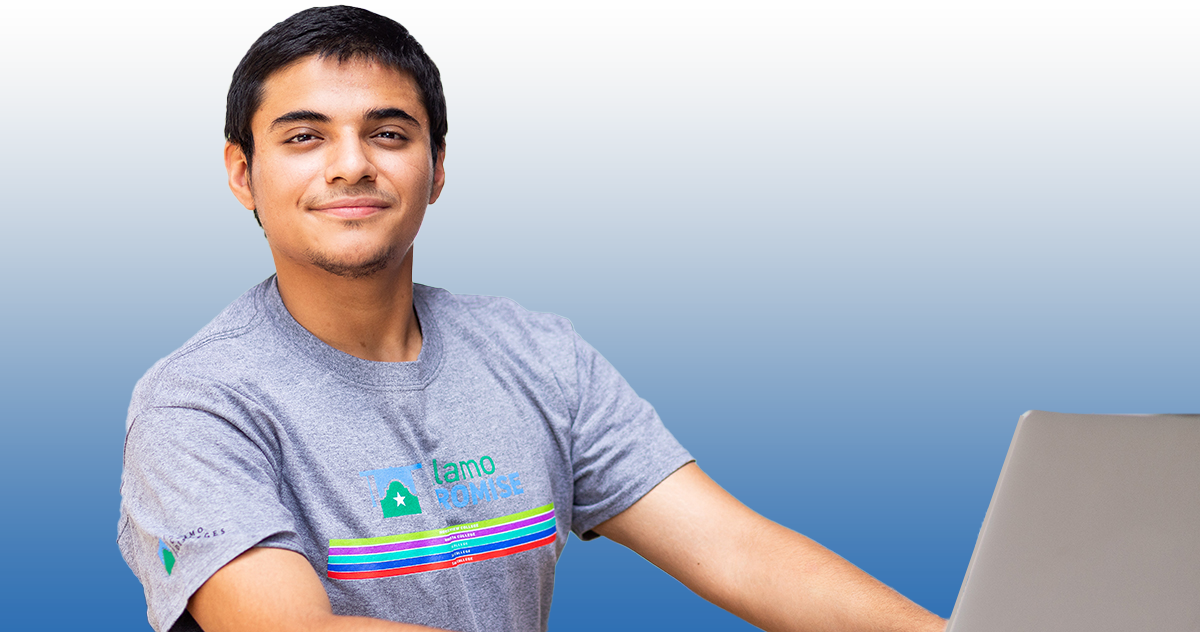 男学生在笔记本电脑上工作时微笑;一名学生穿着一件前面印有阿拉莫承诺标志的灰色衬衫